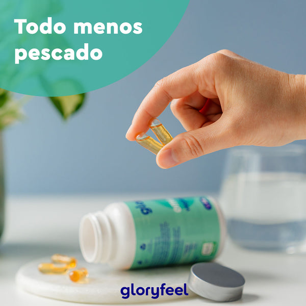 Una mano sostiene una cápsula sobre una botella derramada de Omega 3 Vegano en cápsulas de GloryFeel GmbH España sobre una mesa. El texto dice "Todo menos pescado". Al fondo, un vaso de agua y los suplementos de Omega 3.
