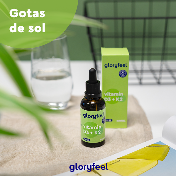 Sobre una mesa, junto con un vaso de agua, hay un frasco con gotero y una caja de Vitamina D3/K2 en gotas de GloryFeel GmbH España. El texto dice: «Gotas de sol para tu salud ósea».