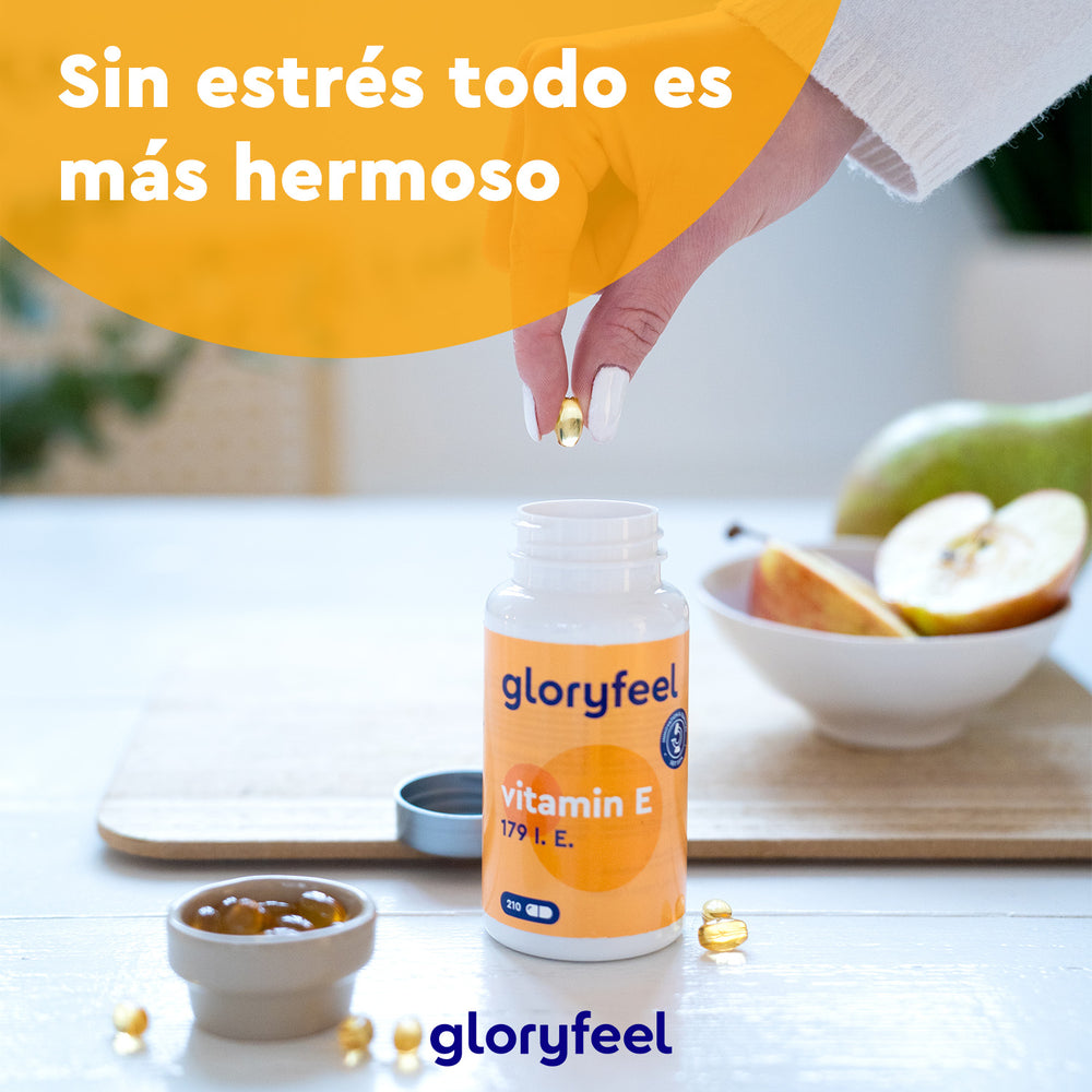 Una mano sostiene una cápsula de vitamina E de 179 UI sobre una botella abierta de GloryFeel GmbH España, rodeada de fruta y un bol. Texto en español: «Con tocoferoles, sin estrés, todo es más hermoso».
