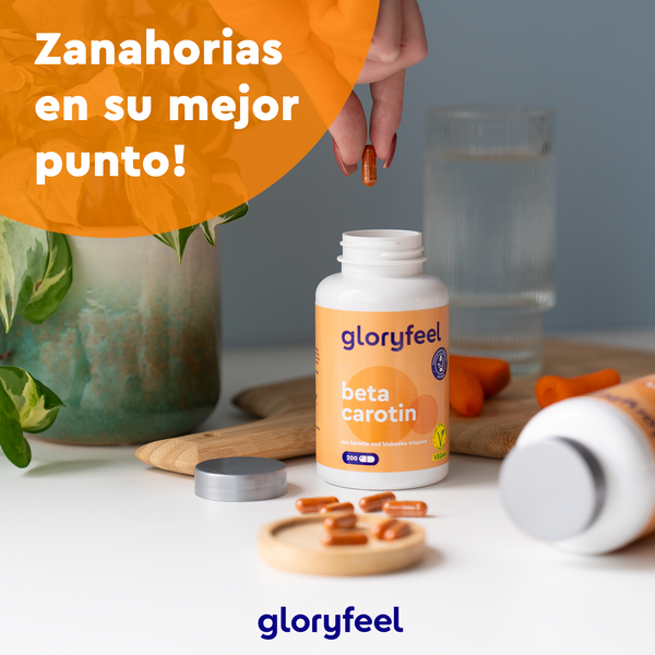 Una botella de Betacaroteno de Gloryfeel GmbH está rodeada de zanahorias frescas y un vaso de agua. Un círculo naranja con el texto en español &quot;¡Zanahorias en su mejor punto!&quot;, destacando los potentes carotenoides para tu aporte diario de vitamina A.