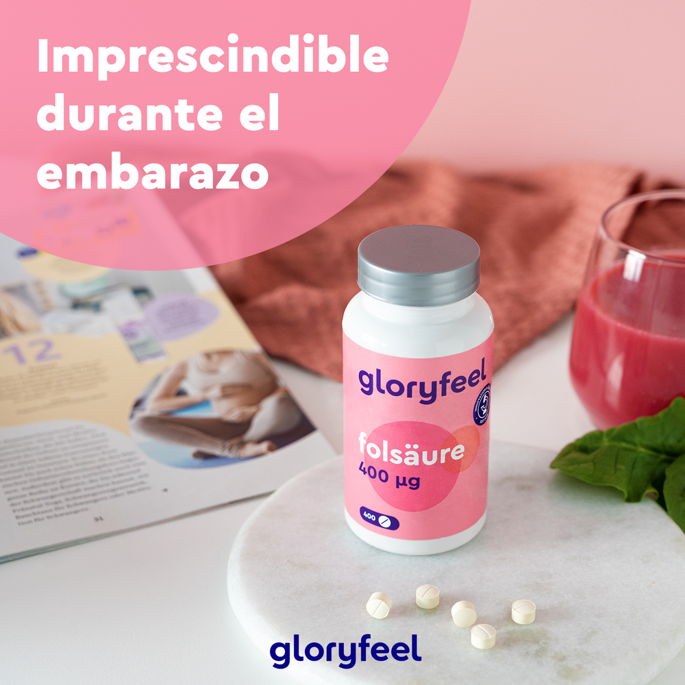 Un frasco de Ácido Fólico (Vitamina B9) de Gloryfeel GmbH está sobre una mesa con pastillas de folato, un libro, una bebida rosa y espinacas. El texto dice &quot;Imprescindible durante el embarazo&quot;, lo que enfatiza la importancia de estas pastillas fáciles de tragar.