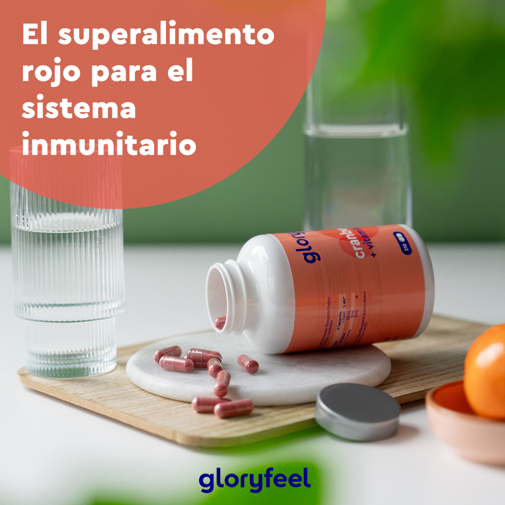 Una botella abierta de cápsulas de Arándano Rojo + Vitamina C de Gloryfeel GmbH se encuentra junto a pastillas esparcidas, un vaso de agua, una naranja y hojas de plantas. El texto dice: &quot;El superalimento rojo para el sistema inmunológico.