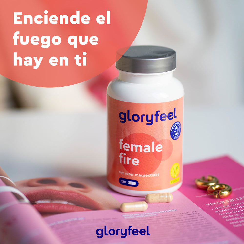 Una botella de &quot;Female Fire: con Maca Roja&quot; de GloryFeel GmbH España reposa sobre una revista abierta, rodeada de cápsulas. El texto en español dice: &quot;Enciende el fuego que hay en ti&quot;.