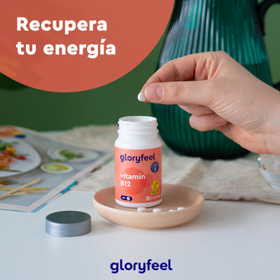 Una mano toma una tableta de Vitamina B12 en tabletas de GloryFeel GmbH Spain de un frasco abierto sobre un plato, con el texto "Recupera tu energía" y resaltando cómo la vitamina apoya tus glóbulos rojos.