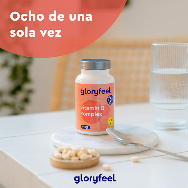 Una botella de Tabletas del complejo de vitamina B de GloryFeel GmbH España reposa sobre un posavasos de mármol con un vaso de agua y un plato de pastillas. Al fondo, una silla y plantas, con el texto &quot;Ocho de una sola vez&quot;, que destaca el bienestar en cada dosis.