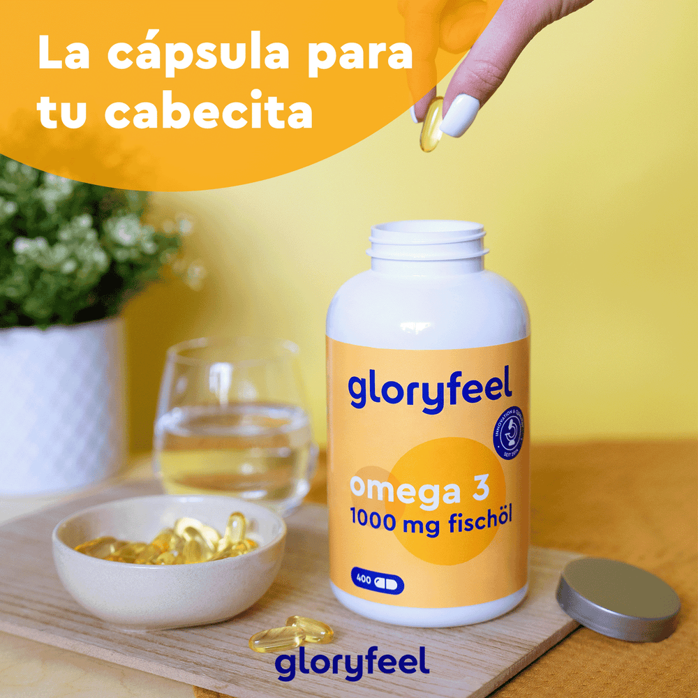 Una mano sostiene con cuidado una cápsula de omega-3 sobre un frasco con la etiqueta &quot;Omega 3 en cápsulas&quot; de GloryFeel GmbH España, junto a un vaso de agua y un bol con suplementos alimenticios. El texto de la imagen está en español.