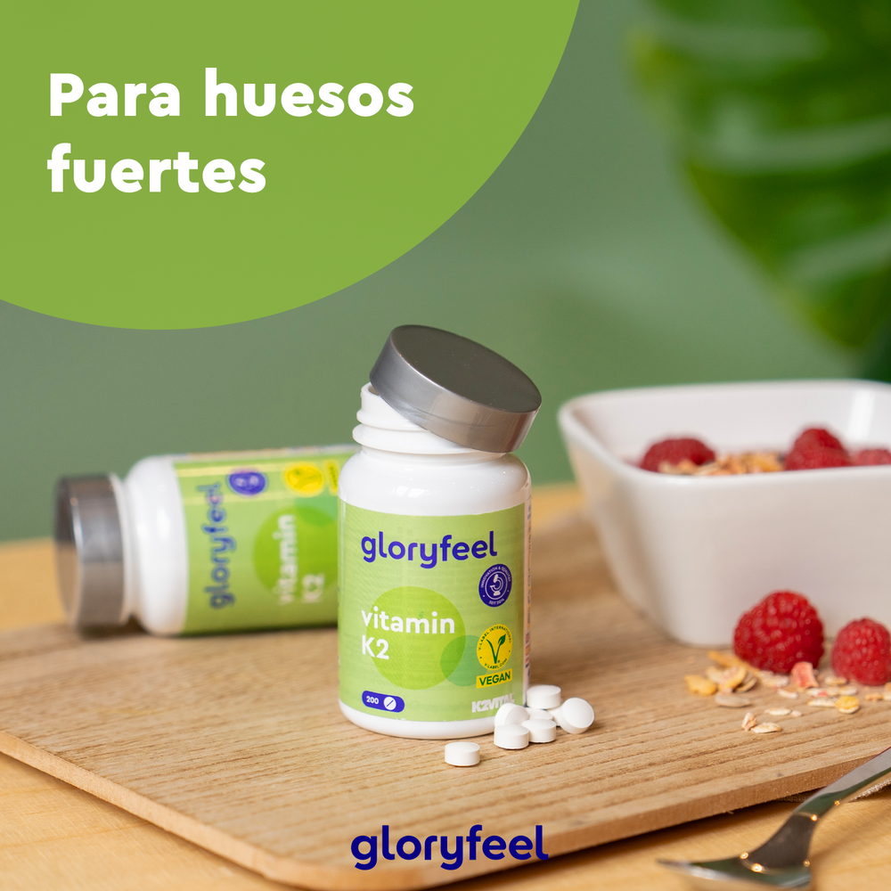 Dos frascos de vitamina K2 en tabletas de gloryfeel GmbH están sobre una tabla de madera con una pequeña pila de tabletas. Al fondo, un tazón de cereales con frambuesas. El texto dice: &quot;Para huesos fuertes con menaquinona&quot;.