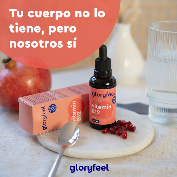 Una botella de Vitamina B12 en Gotas de GloryFeel GmbH reposa sobre una mesa rodeada de semillas de granada, una cuchara y un vaso de agua. El texto en español dice: «Tu cuerpo no lo tiene, pero nosotros sí», ideal para veganos y vegetarianos.