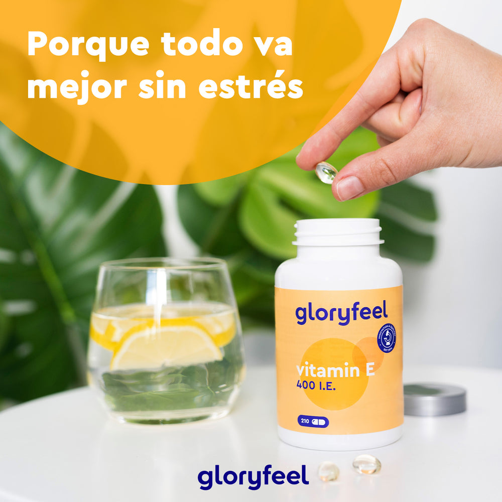 Una mano sostiene una cápsula sobre un frasco de vitamina E de GloryFeel GmbH, conocido por su contenido en tocoferoles, junto a agua con limón. El texto dice: &quot;Porque todo va mejor sin estrés&quot;, con una planta exuberante que le da un toque antioxidante de fondo.