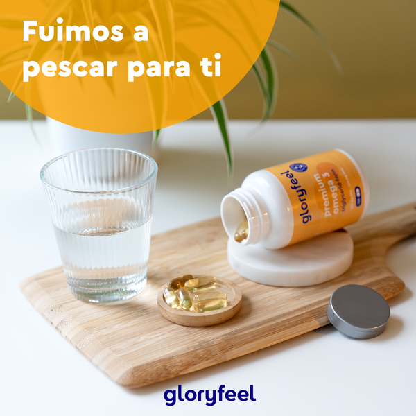 Una botella de Omega 3 en forma de triglicéridos de GloryFeel GmbH España, rico en EPA y DHA, descansa sobre una superficie de madera junto a un vaso de agua y un pequeño plato, con un texto en español que dice: &quot;Fuimos a pescar para ti.