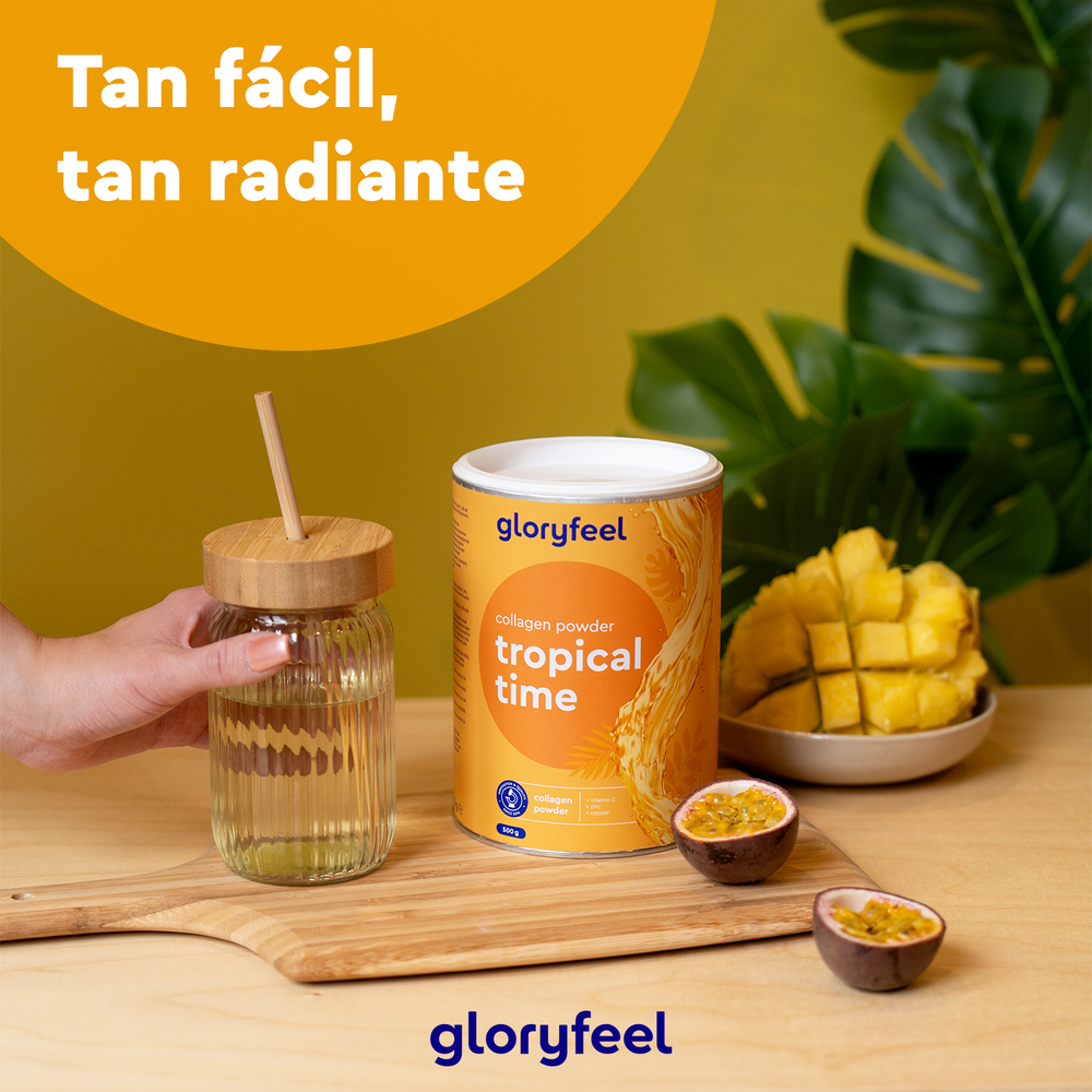 Sobre una mesa, hay una lata de Colágeno en polvo de 500 g - Tropical time - Sabor a frutas tropicales junto a un frasco de vidrio, maracuyá en rodajas y mango picado. Al fondo, hojas verdes. El texto dice &quot;Tan fácil, tan radiante&quot; junto a &quot;GloryFeel GmbH Spain&quot; y contiene vitamina C añadida para una luminosidad extra.
