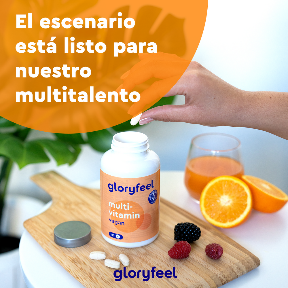 Una mano toma una pastilla de un frasco de GloryFeel GmbH Spain Multivitaminas veganas en tabletas sobre una tabla de cortar, rodeada de bayas, rodajas de naranja y un vaso de zumo de naranja. El texto en español dice: «El escenario está listo para nuestro multitalento de antioxidantes».