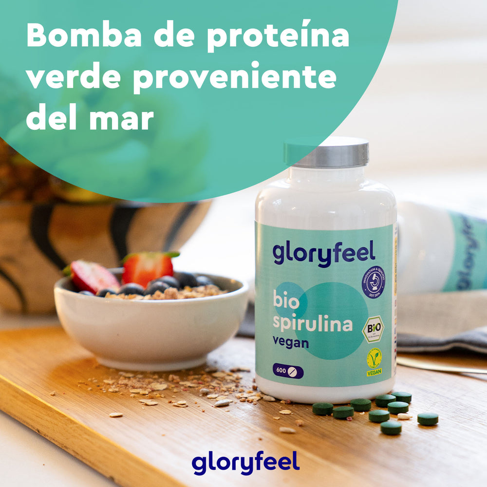 Una botella de GloryFeel GmbH España Espirulina Ecológica - Tabletas se encuentra sobre una mesa junto a un tazón de cereales cubierto con fresas y arándanos, ricos en nutrientes y vitaminas.
