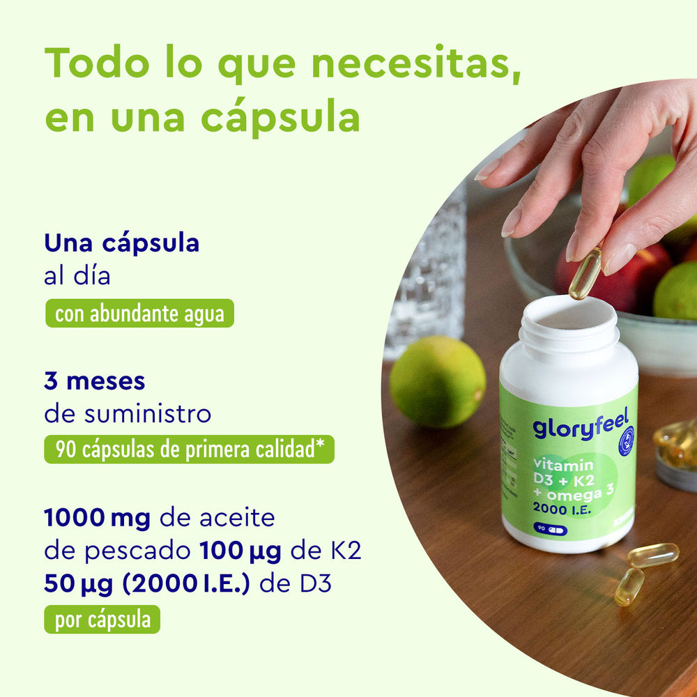 Una mano sostiene una cápsula junto a una botella de GloryFeel GmbH etiquetada &quot;Vitamina D3/K2 &amp; Omega 3 2000 UI&quot; sobre una mesa con detalles de dosis e ingredientes en español, acompañada de frutas y un vaso de agua en el fondo.