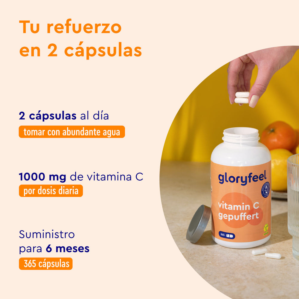 Sobre una mesa hay una botella abierta de Cápsulas de Vitamina C Tamponada de GloryFeel GmbH España, con una mano sosteniendo dos cápsulas encima. La dosis y los detalles del suplemento están resaltados en texto, con frutas visibles al fondo.