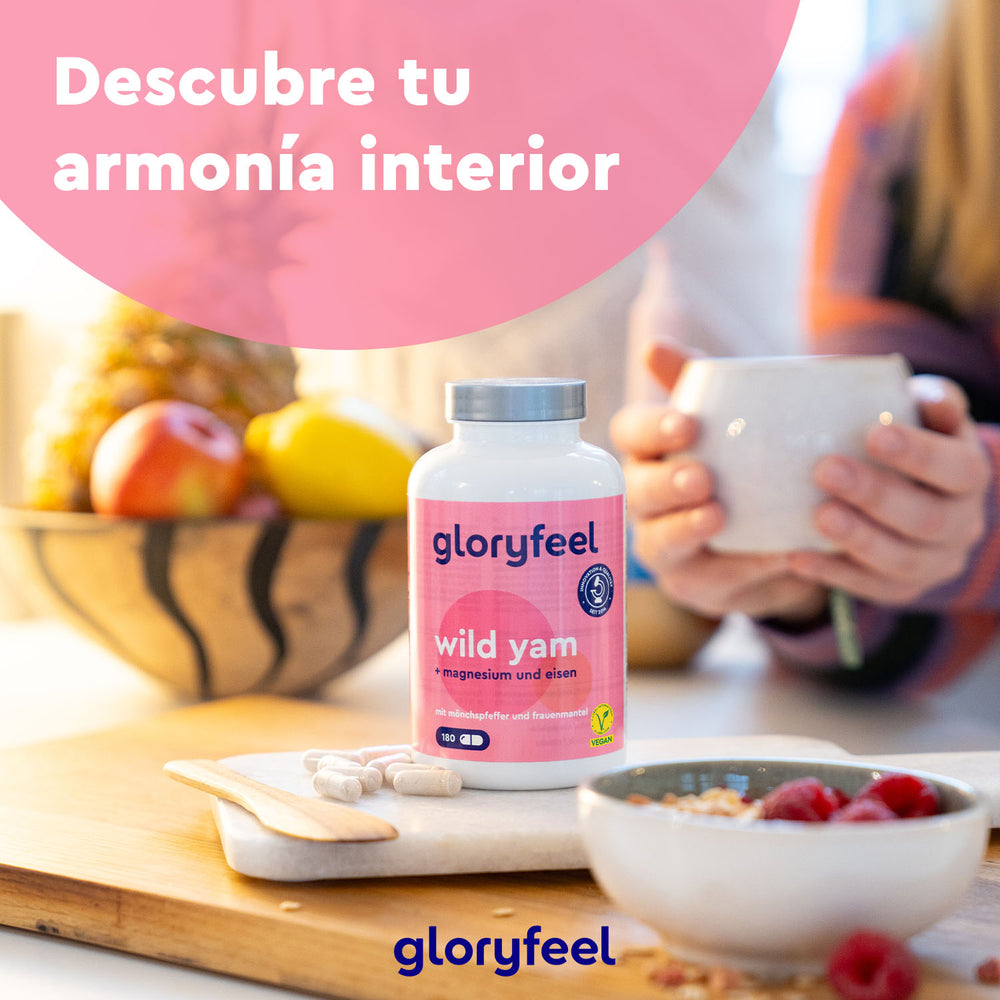 Una botella de Wild Yam o raíz de ñame silvestre, conocida por la diosgenina, de Gloryfeel GmbH reposa sobre una mesa con fruta, mientras alguien en el fondo sostiene una taza.